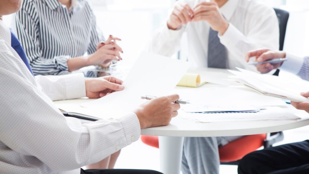 テーブルを囲んで行われている会議で、資料やペンを手に持っているビジネスパーソンたちの手元のアップ