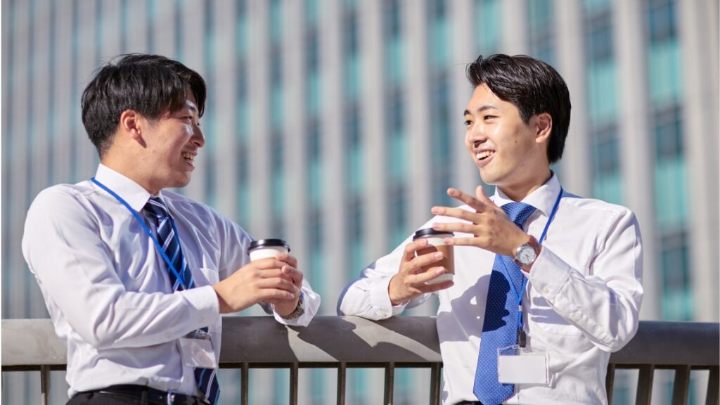屋外で紙コップのコーヒーを手に持ち、笑顔で談笑している2人の若い男性ビジネスマン