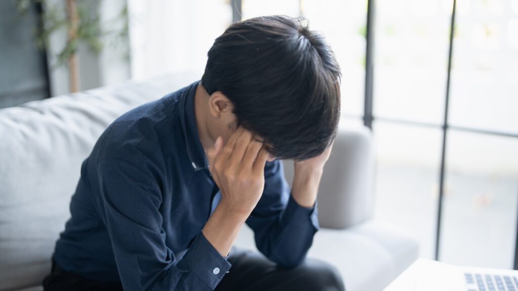 ソファに座り、両手で頭を抱えてうつむいて悩んでいる若い男性
